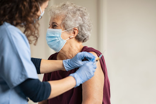 A senior woman receives a COVID-19 vaccine shot. She is wearing a mask. A female nurse is administering the dose.